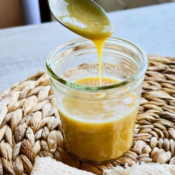 Thick golden vegan sweetened condensed milk drips from a spoon into a glass jar on a woven placemat.
