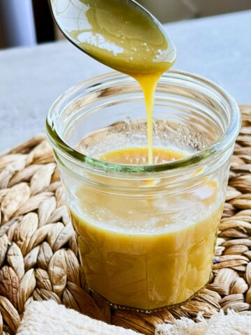 Thick golden vegan sweetened condensed milk drips from a spoon into a glass jar on a woven placemat.