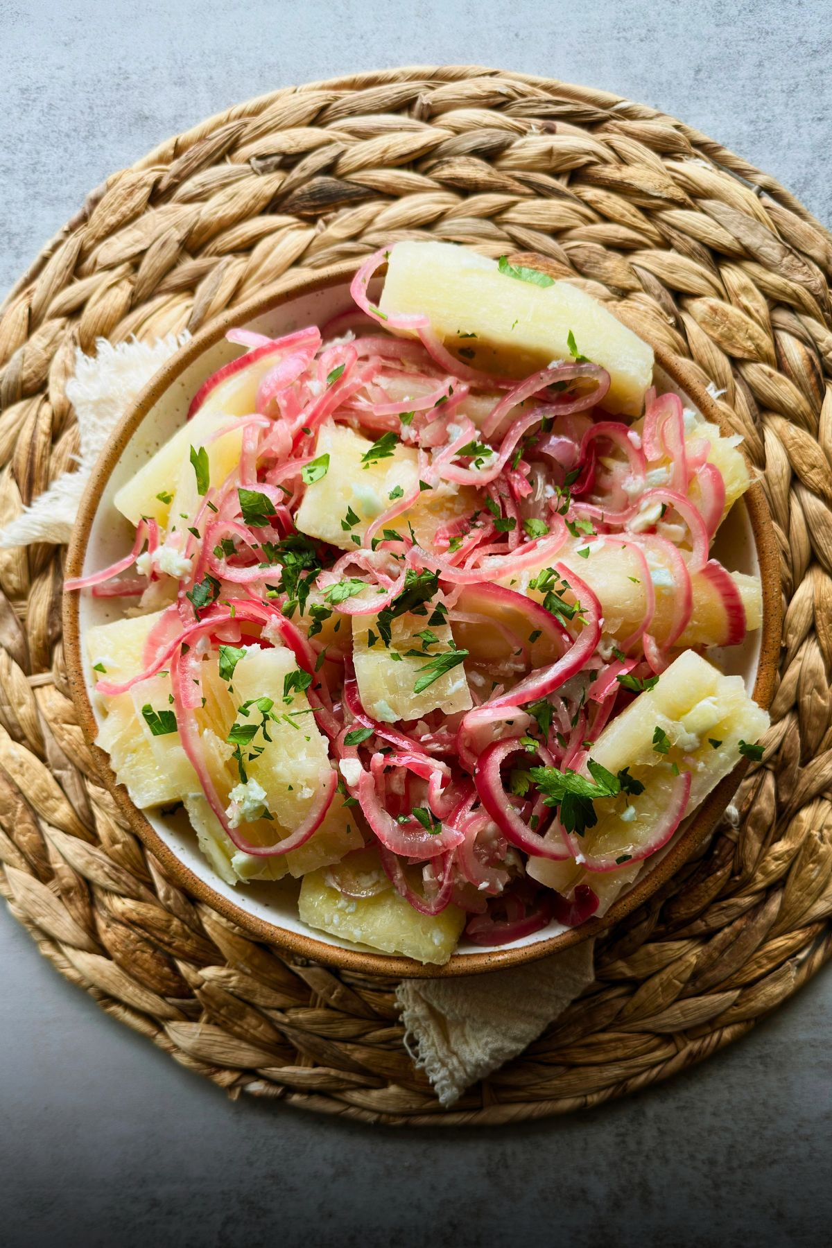 Yuca con mojo topped with pink onions and parsley in a bowl on a woven placemat.